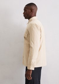 Beżowa bawełniana koszula z długim rękawem, wyposażona w kieszeń na piersi, mankiety na guziki oraz klasyczny kołnierzyk, noszona na ciemnych spodniach.