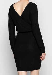 Robe en maille noire avec un dos en V profond, des manches longues et une taille cintrée. La texture est côtelée, présentant une silhouette élégante et moulante.