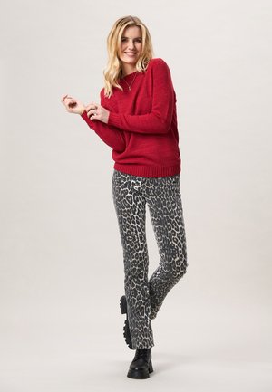 Vrouw met blond haar, gekleed in een rode trui, broek met luipaardprint en zwarte laarzen, staand met één been licht gekruist.