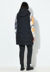 Svart vadderad väst med huva, med en lång silhuett, kombinerad med en flerfärgad mönstrad skjorta med långa ärmar och mörka jeans.