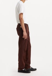 Pantalones de algodón marrón con corte recto, cinco bolsillos y una etiqueta en el bolsillo trasero, combinados con una camiseta blanca lisa y zapatos negros.