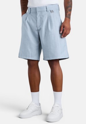 Mann trägt hellblaue knielange Shorts, weiße Turnschuhe, weiße Sportsocken und ein weißes ärmelloses Shirt vor einfarbigem Hintergrund.