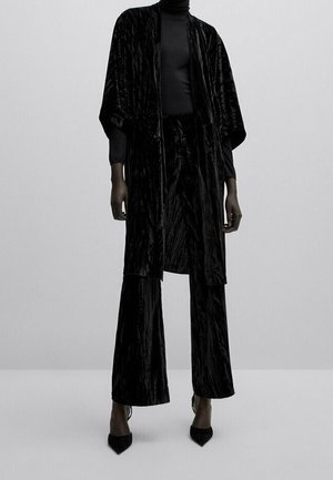 Persona che indossa una giacca in stile kimono di velluto nero, pantaloni a gamba larga coordinati, dolcevita nero e tacchi neri con punta appuntita, in piedi su pavimento grigio.