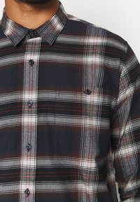 Granatowa koszula w kratę z czerwonymi i białymi paskami, z kołnierzykiem typu spread i czarnymi guzikami, wykonana z miękkiego materiału.