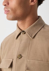 Veste en daim beige avec un col pointu, deux poches poitrine et une fermeture à boutons avec un design circulaire et quatre trous.