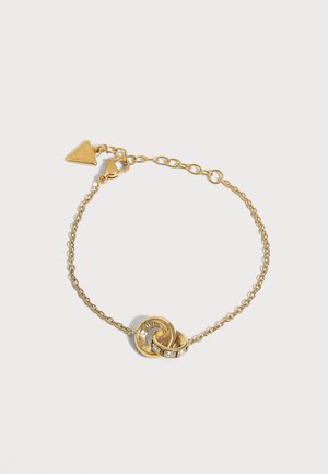 Bracelet délicat en chaîne d’or avec deux anneaux entrelacés, l’un gravé et l’autre serti de petites pierres claires, et un pendentif en forme de cœur près du fermoir.