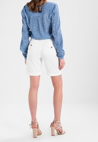 Donna con una blusa a maniche lunghe di un blu chiaro, pantaloni corti bianchi fino al ginocchio con tasche posteriori e sandali con tacco beige, in piedi di spalle.