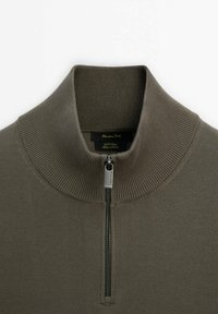 Dunkelolivgrüner Pullover mit hohem Kragen, ausgestattet mit einem Metallreißverschluss und Rippstruktur. Enthält ein Markenetikett im Halsbereich.