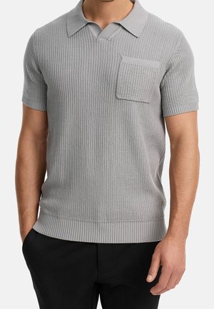 Homme portant un polo en maille à manches courtes gris clair avec une poche poitrine et un pantalon noir, debout la main dans la poche.