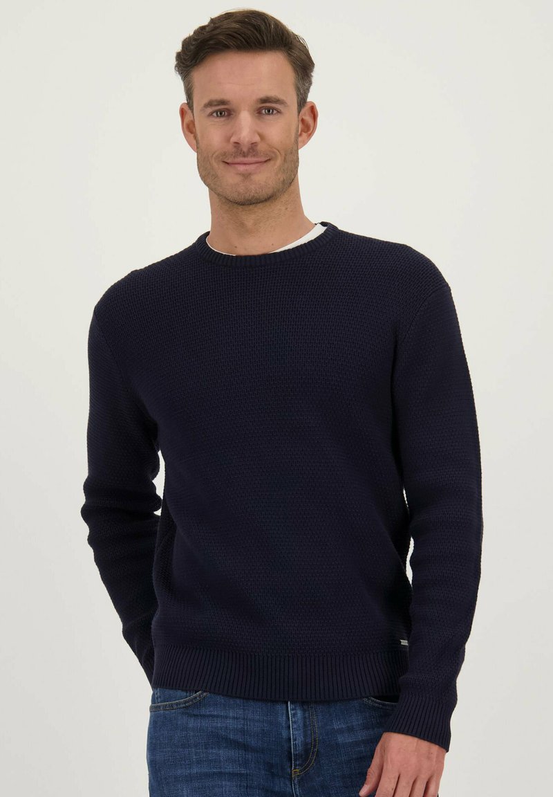 Marineblauwe gebreide trui met textuur, ronde halslijn en geribde boorden en zoom, gedragen over een wit shirt. Casual pasvorm en veelzijdig ontwerp.