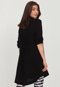 Schwarze Tunika mit lockerer Passform, kurzen puffigen Ärmeln, gerafftem Rückendetail und asymmetrischem Saum, kombiniert mit gemusterten Leggings.