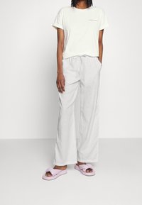 Personne portant un t-shirt blanc avec le texte "Colourful Rebel", un pantalon ample gris clair avec cordon de serrage, et des sandales glissantes violettes claires.