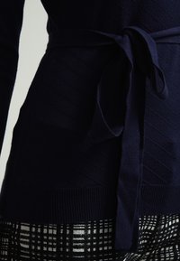 Haut tricoté marine avec un motif diagonal texturé. Comprend une ceinture nouée à la taille et un ourlet côtelé. Associé à un pantalon à motif écossais.