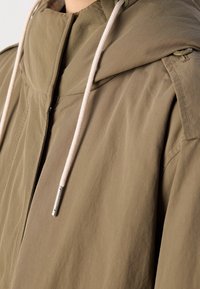Giacca con cappuccio verde oliva con chiusura a zip, in tessuto leggero, cordini beige regolabili e dettagli di cucitura discreti sulle spalle.