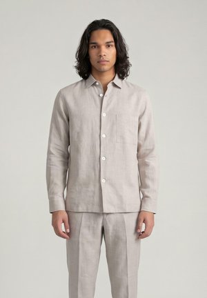 Man met halflang donker haar die een beige overhemd met lange mouwen en bijpassende broek draagt, staand tegen een effen achtergrond.