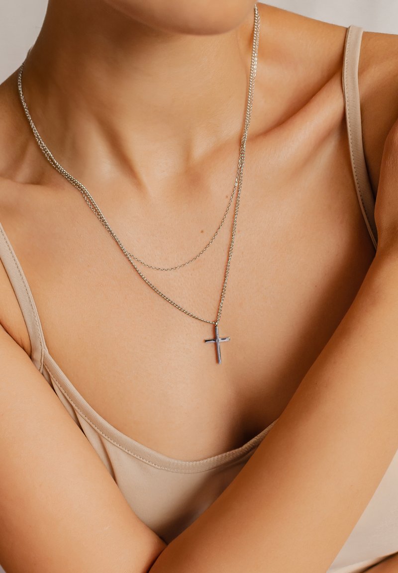 Collier en croix argenté avec double chaîne, présentant une texture lisse et un design minimaliste. Porté par-dessus un haut ajusté et léger.