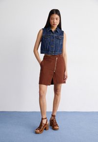 Denim ermeløs vest, mørkeblå, med frontlommer. Brun skjørt med frontglidelås og splitt. Brune hæl-sandaler med stropper.