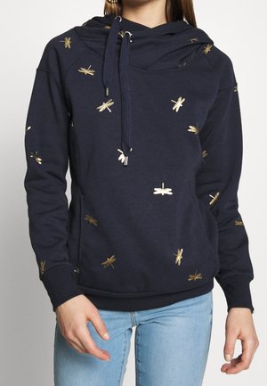 Kobieta w granatowej bluzie z kapturem z nadrukiem złotych ważek, w połączeniu z jasnoniebieskimi jeansami, stojąca na jednolitym tle.