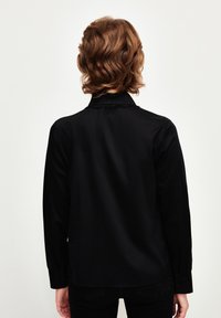 Maglietta nera a maniche lunghe con colletto e polsini abbottonati. Tessuto liscio, vestibilità rilassata e delicate cuciture nella parte posteriore.