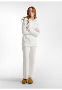 Sudadera blanca y suave con un bolsillo frontal, combinada con pantalones tapered a juego. Modelo lleva zapatillas de felpa tipo pantuflas en color beige.