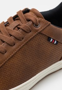 Sneakers en cuir marron avec une empeigne perforée, ornées de lacets beiges et d'une petite étiquette en rouge, blanc et bleu. Semelle en caoutchouc blanche.