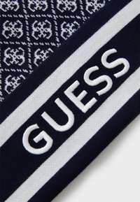 Strikket skjerf med marineblå bunn, med hvite geometriske mønstre og en fremtredende hvit "GUESS" logo i brodert tekst.