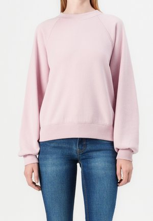 Žena v světle růžové dlouhém mikině s žebrovanými manžetami a modrých džínách stojí na bílém pozadí.