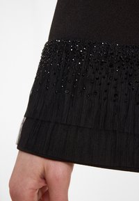 Schwarzes Kleid mit Ärmeln, das ein strukturiertes Design mit gestuften Rüschen aufweist und am Saum mit schwarzen Pailletten und Perlen verziert ist.