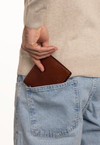 Brown leather wallet partially visible in a blue denim pocket, held by a hand. The wallet features a smooth texture and simple design.