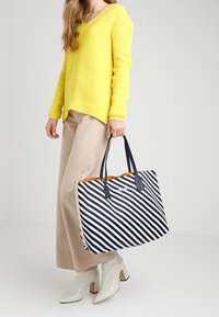 Donna che indossa un maglione giallo e pantaloni beige tiene in mano una grande borsa a tracolla a righe navy e bianche con manici neri.