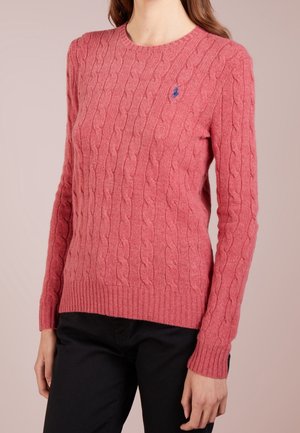 Vrouw draagt een roze kabelgebreide trui met een kleine blauwe geborduurde logo op de borst en een zwarte broek.