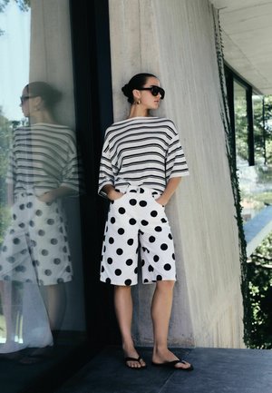 Woman wearing black sunglasses, striped white and black top, and white shorts with large black polka dots, standing against a concrete wall.