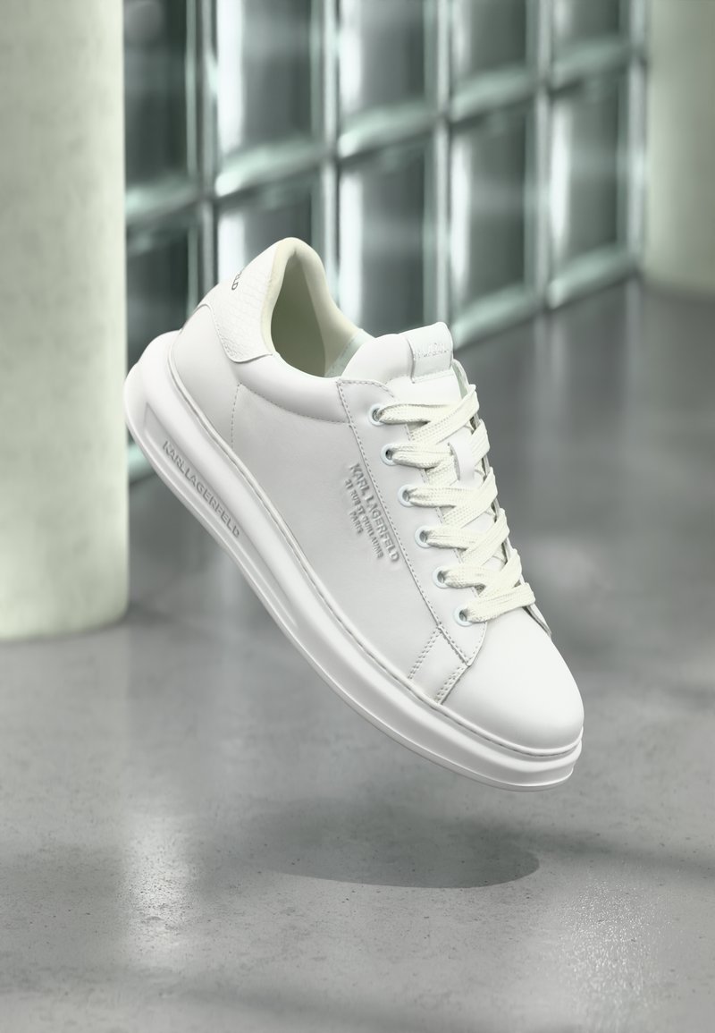 Fehér Karl Lagerfeld sneaker fűzőkkel, emboszált logóval az oldalon és a saroknál, felfüggesztve a szürke padló fölött egy modern, üvegfalú szobában.