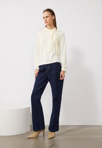 Cremfarbene Bluse mit langen Ärmeln und gebundenem Halsdetails, kombiniert mit dunkelblauen geraden Jeans. Hellbeige Knöchelstiefel ergänzen das Outfit.