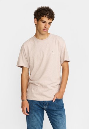 Jonge man met krullend haar, gekleed in een licht beige t-shirt met een kleine geborduurde figuur en een blauwe spijkerbroek, poseert met één hand in de zak.