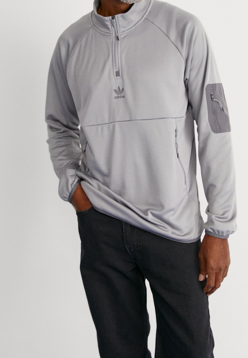 Grå sweatshirtjacka med halvmaskerad krage, strukturerad tyg, Adidas-logotyp och ficka med dragkedja på vänster arm. Kombinerad med mörka jeans.