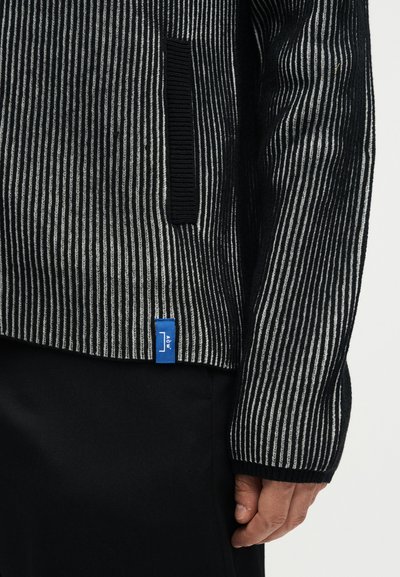 Κοντινό πλάνο ενός ατόμου που φοράει μαύρο και γκρι κάθετα ριγέ πουλόβερ με μια μικρή μπλε ετικέτα κοντά στο τελείωμα και μαύρο παντελόνι.