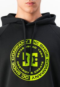 Close-up of a person wearing a black hoodie with a neon green circular DC Shoe Company logo and drawstrings hanging down.