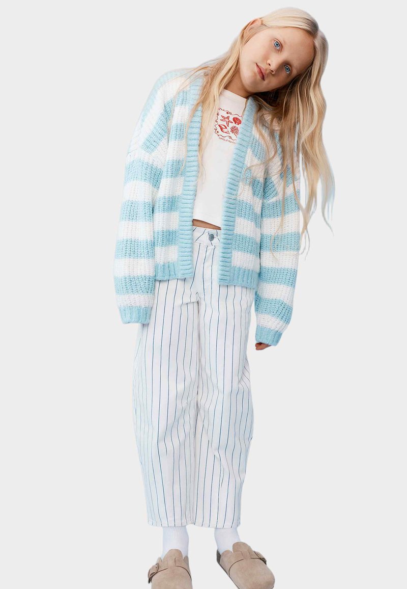 Niña joven con cabello largo y rubio que lleva un cárdigan a rayas azul y blanco, camiseta blanca con diseño gráfico, pantalones de rayas diplomáticas y zapatos marrones sin cordones.