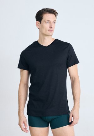 Homme avec des cheveux bruns courts portant un T-shirt noir à col en V et un caleçon vert foncé, regardant vers sa droite sur un fond uniforme clair.