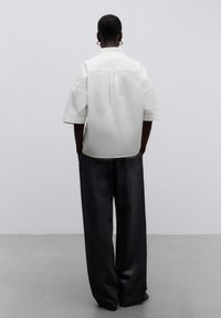 Top bianco a maniche corte con collo alto e spacco sul retro, abbinato a pantaloni larghi e scuri a gamba larga, in tessuto liscio e design minimalista.