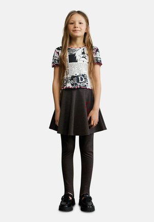 Jong meisje met lang haar, gekleed in een zwart-wit grafisch T-shirt, een donker gestreepte rok en legging, en zwarte loafers, staat recht vooruit gericht.