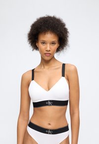 Νεαρή γυναίκα με σγουρά μαλλιά, φοράει λευκό σουτιέν και εσώρουχο Calvin Klein με μαύρους ιμάντες και λάστιχο μέσης, σε απλό φόντο.