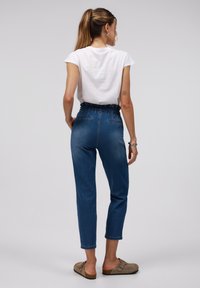 Lichtblauwe spijkerbroek met een hoge taille en elastische tailleband. Beschikt over taps toelopende benen en achterzakken. Gecombineerd met een wit T-shirt en beige instappers.