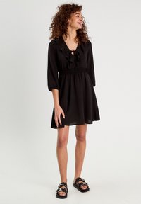 Robe noire avec un col froncé, des manches trois-quarts et une taille élastiquée. Caractérisée par une jupe évasée. Portée avec des sandales noires à clous.