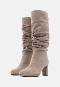 Beige zamša augstpapēžu zābaki ar brīvu dizainu, apaļu purngalu un masīlu papēdi. Tekstūra ir maiga, ar sakropļotu materiālu ap augšējo daļu.