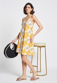 Vestido amarillo y blanco con patrones y cuello halter, que presenta motivos florales y un diseño de texto en contraste, combinado con un bolso de malla negro.