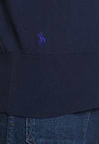 Acercamiento al dobladillo de un suéter de punto azul marino con un logo de jugador de polo bordado en púrpura, justo encima del bolsillo de unos jeans de mezclilla azules.