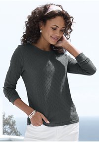 Dunkelgraues Langarmoberteil mit einem strukturierten Rautenmuster, Rundhals und enganliegenden Ärmeln. Kombiniert mit weißen Hosen und silbernen Armbändern.