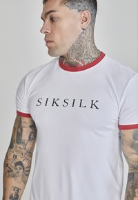 Witte fitted t-shirt met rode geribbelde kraag en mouwaccenten, met de tekst "SIKSILK" in zwart. Model heeft tatoeages zichtbaar op zijn armen en hals.